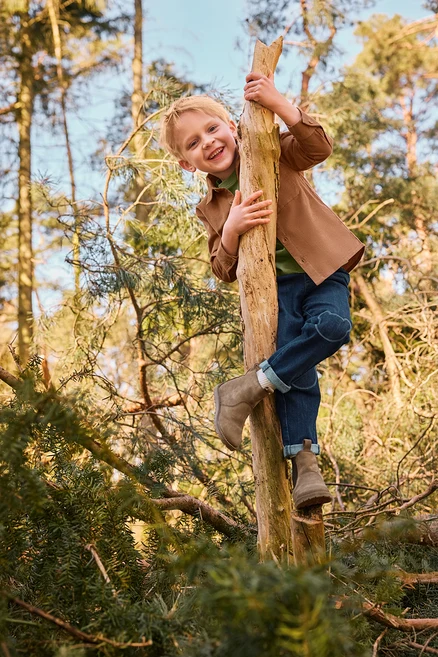 Usměvavý chlapec leze na kmen stromu v lese, oblečený v hnědé bundě, zeleném tričku, modrých džínách s nášivkami na kolenou a světle hnědých botách.