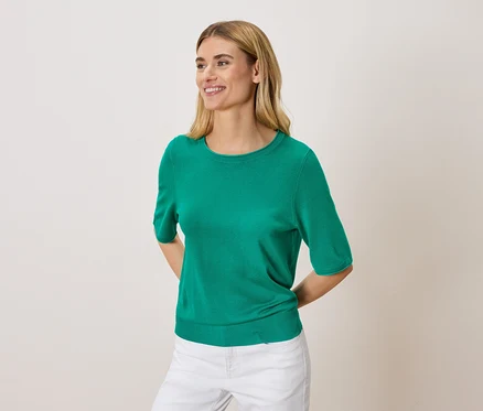Žena s blond vlasy má na sobě zelený svetr a bílé kalhoty.