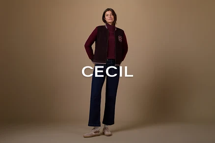 Žena stojí s rukama v kapsách, oblečená v tmavě červeném roláku, tmavě hnědé bomber bundě s bílou nášivkou „B“, tmavě modrých rozšířených džínách a světle růžových teniskách s bílými ponožkami, na jednobarevném hnědém pozadí.