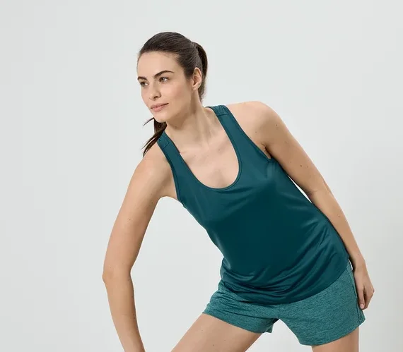 Žena ve sportovním oblečení, skládajícím se ze zeleného tílka a šortek, se protahuje na světlém pozadí.
