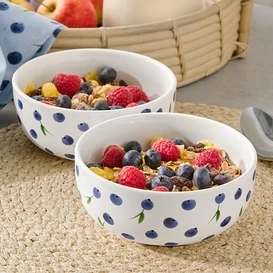 Dvě misky s müsli, malinami a borůvkami na prostírání. V pozadí džbán s mlékem a jablka v košíku.