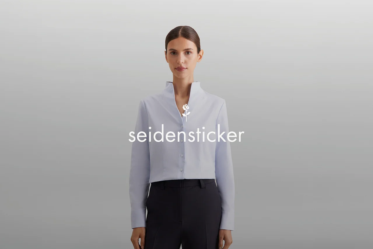 Žena s vlasy staženými dozadu má na sobě světle modrou zkrácenou košili s vysokým límcem a tmavé kalhoty. Na hrudi je bílé logo „seidensticker“ s růží.