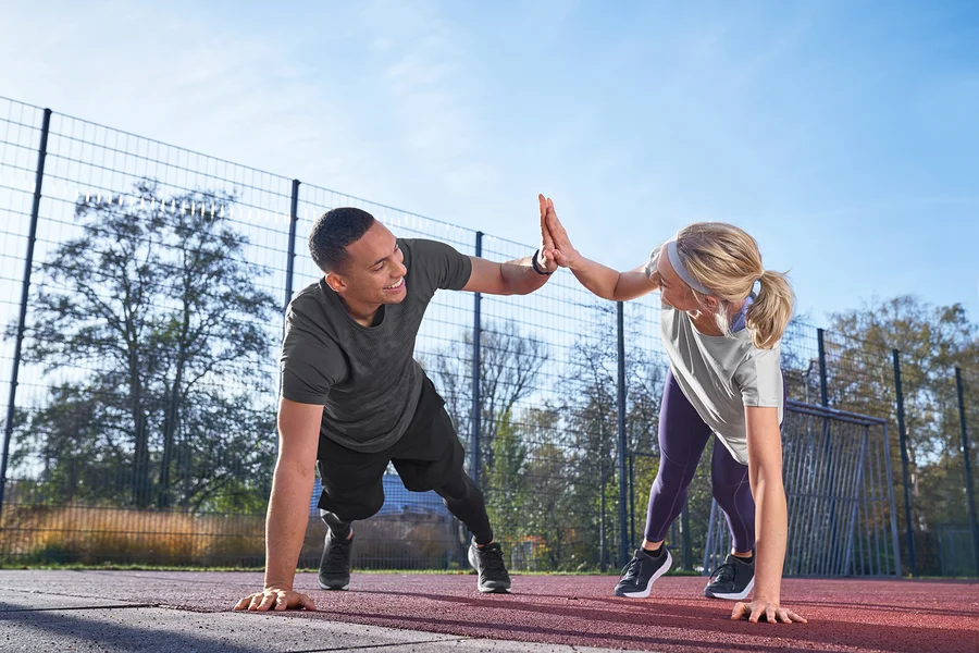 Muž ve sportovním tričku světle šedé s melírem a sportovních šortkách a žena v legínách a funkčním tričku Klima-Tec si plácají rukama při cvičení venku.