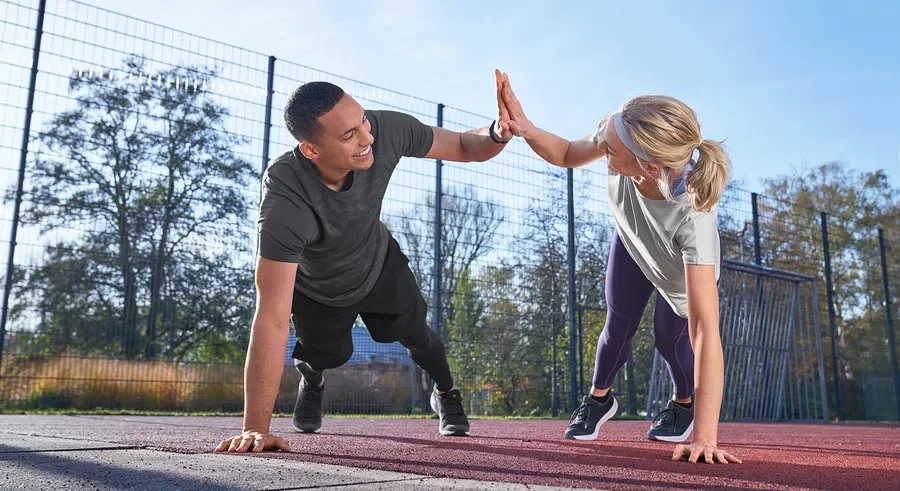 Muž ve sportovním tričku světle šedé s melírem a sportovních šortkách a žena v legínách a funkčním tričku Klima-Tec si plácají rukama při cvičení venku.