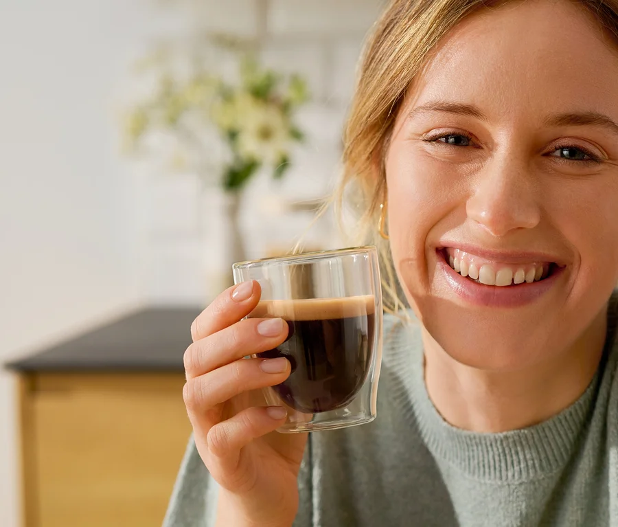 Usmívající se žena drží sklenici Cafissimo Flavoured Espresso – Toasted Nut vedle kávovaru Cafissimo pure+, white.