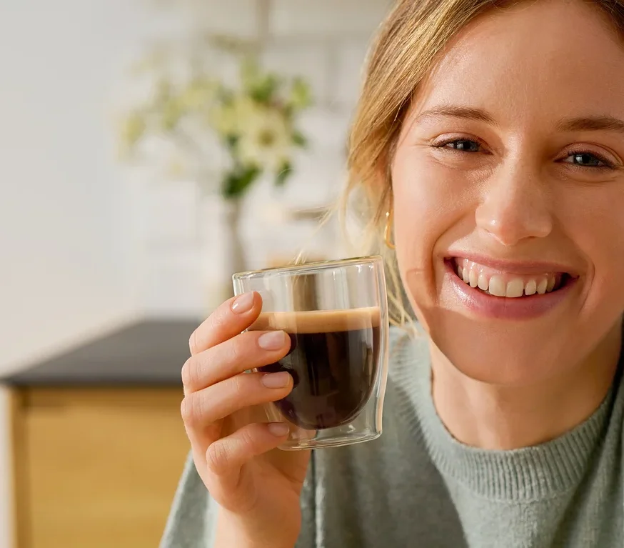 Usmívající se žena drží sklenici Cafissimo Flavoured Espresso – Toasted Nut vedle kávovaru Cafissimo pure+, white.