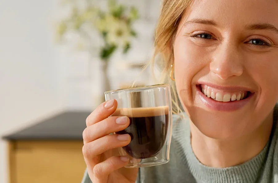 Usmívající se žena drží sklenici Cafissimo Flavoured Espresso – Toasted Nut vedle kávovaru Cafissimo pure+, white.