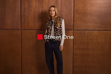 Žena s dlouhými blond vlasy stojí před dřevěnou stěnou, oblečená v leopardí vestě přes bílý top s dlouhým rukávem a tmavé džíny. Má ruce v kapsách. Viditelné je logo "Street One".
