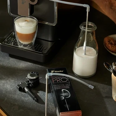 Antracitový kávovar Tchibo »Esperto Pro« pripravuje nápoj. Mlieko prechádza trubicou do sklenenej fľaše. Vedľa je náhradná sada pre Esperto Pro a Esperto2 Milk.