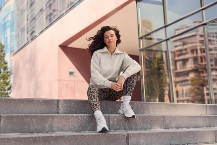 Žena s kudrnatými vlasy sedí na betonových schodech, má na sobě béžový top s polovičním zipem, legíny s leopardím vzorem a bílé tenisky, v pozadí jsou moderní budovy.