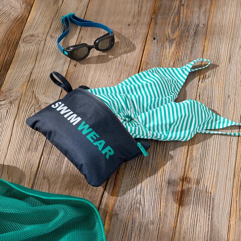 Snímek plaveckých doplňků na dřevěném povrchu včetně tašky s nápisem „Swimwear“, zeleno-bílých pruhovaných plavek, plaveckých brýlí a zelené síťky.