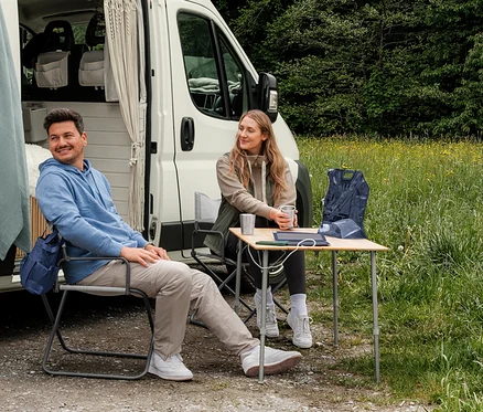 Muž v světle modré mikině s kapucí a žena v béžové mikině a khaki hybridní vestě sedí před karavanem. Na stole leží tmavě modrá sbalitelná vesta.