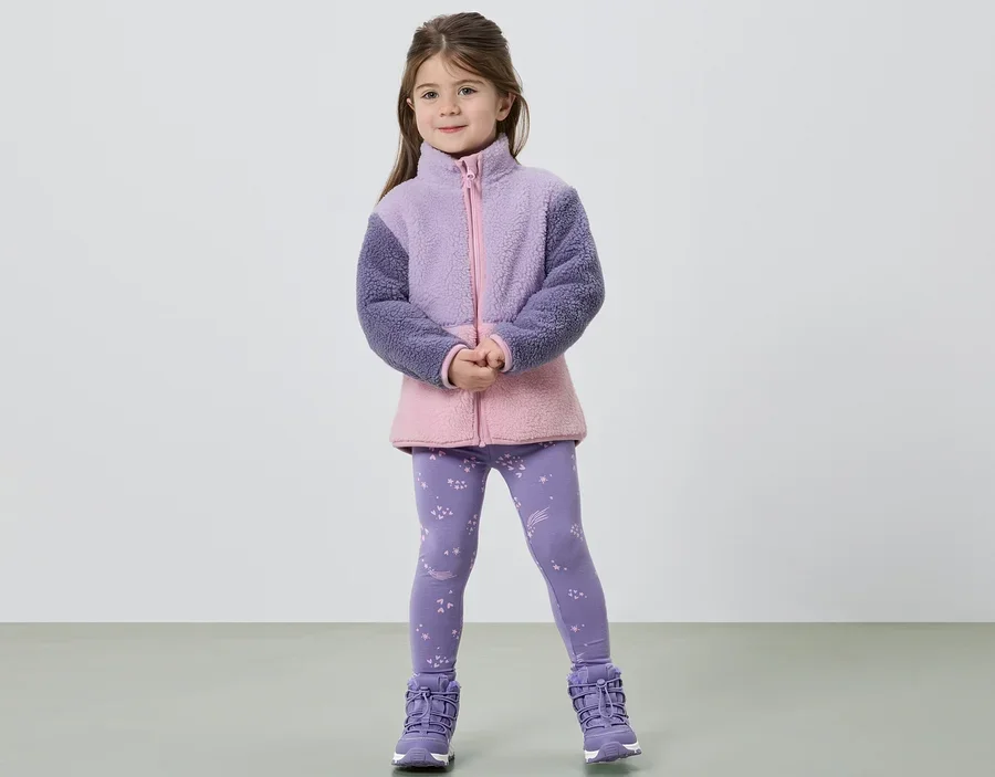 Dívka stojí se sepjatýma rukama a má na sobě fleecovou bundu ve fialové a růžové barvě, legíny s hvězdami a fialové boty.