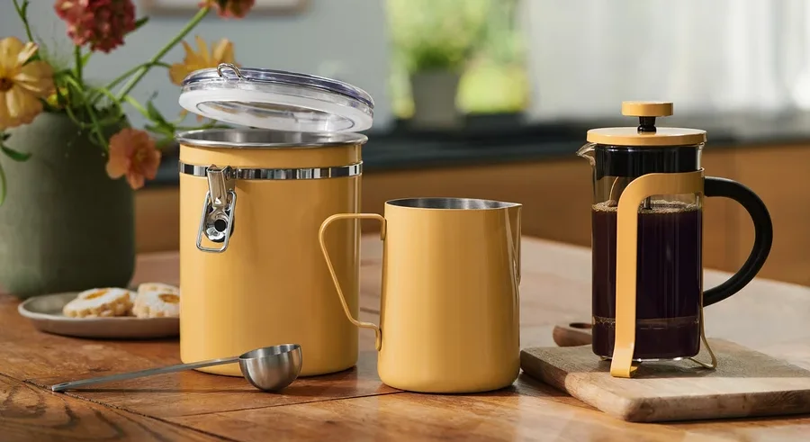Žlutá úložná nádoba s otevřeným víkem, odpovídající hrnek, odměrka a French Press s kávou na dřevěném stole. Květiny a sušenky v pozadí.