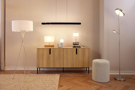 Moderní pokoj s dřevěnou komodou, na které jsou tři lampy a dekorativní předmět, pod černým lineárním závěsným svítidlem. Vlevo na koberci stojí bílá stojací lampa na trojnožce a vpravo vedle šedého taburetu stříbrná stojací lampa s horním světlem a lampou na čtení.