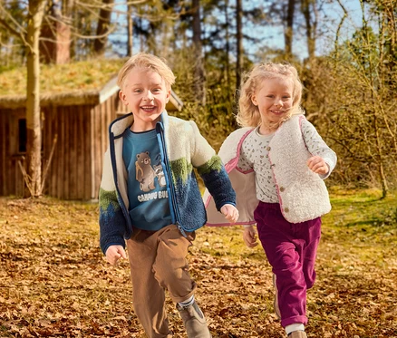Dvě usmívající se děti, chlapec a dívka, běží listnatou venkovní oblastí. Chlapec má na sobě modré tričko s grafikou, barevnou fleecovou bundu a hnědé manšestrové kalhoty. Dívka má na sobě vzorované tričko, krémovou chlupatou vestu a purpurové manšestrové kalhoty. V pozadí je dřevěný dům s travnatou střechou.