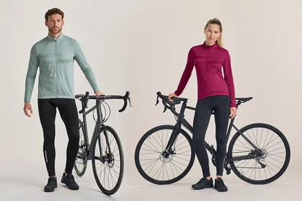 Muž a žena stojí vedle černých silničních kol. Muž má na sobě světle zelený cyklistický dres s dlouhým rukávem a černé cyklistické kalhoty. Žena má na sobě tmavě růžový cyklistický dres s dlouhým rukávem a černé cyklistické kalhoty.