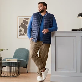 Muž opřený o skříňku má na sobě modrou prošívanou vestu, královsky modrý lněný svetr, kalhoty chino barvy velbloudí srsti a boty sneaker.