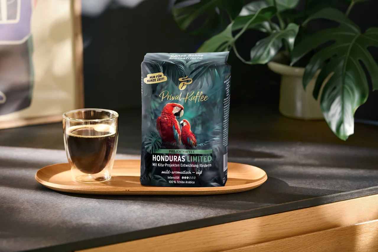 Tmavě zelený sáček kávy „Privat Kaffee Honduras Limited“ se dvěma červenými ary a skleněný šálek černé kávy stojí na dřevěném podnosu. Na sáčku jsou nápisy jako „NUR FÜR KURZE ZEIT!“ a „100% Tchibo Arabica“. V pozadí je zelená rostlina.