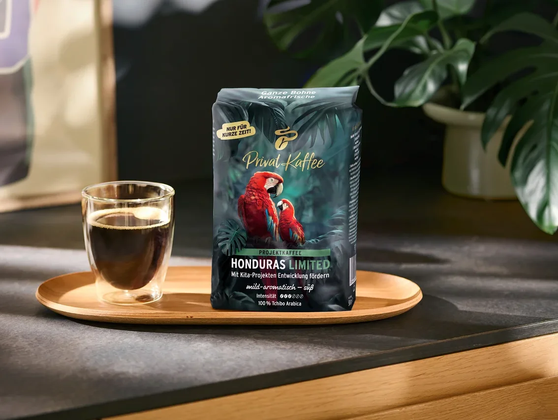 Tmavě zelený sáček kávy „Privat Kaffee Honduras Limited“ se dvěma červenými ary a skleněný šálek černé kávy stojí na dřevěném podnosu. Na sáčku jsou nápisy jako „NUR FÜR KURZE ZEIT!“ a „100% Tchibo Arabica“. V pozadí je zelená rostlina.