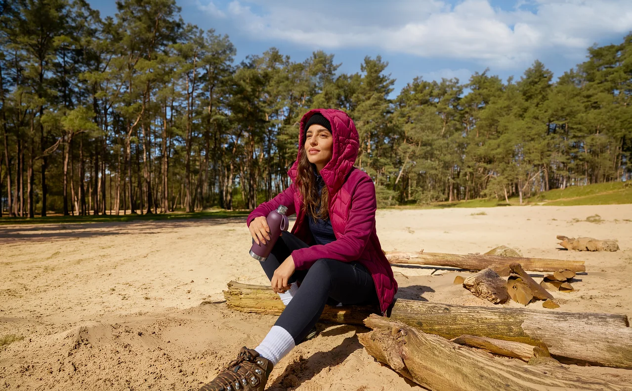 Žena v purpurové bundě sedí na kmeni stromu v písku, drží sportovní láhev na pití a dívá se do nebe.