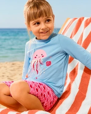 Dívka sedí na lehátku s XL osuškou a má na sobě Dětskou soupravu s UV ochranným faktorem 50+, medúza.