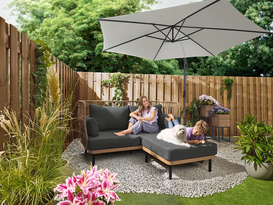 Žena sedí na šedé pohovce pod slunečníkem Leco Ampelschirm Ø 300 cm se stojanem a krytem. Dítě leží se psem na otomane. V pozadí je vidět sada desek Leco pro křížový stojan.