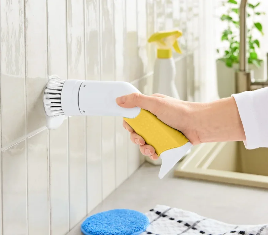 Ruka drží elektrický čisticí kartáč se žlutou rukojetí a bílou hlavicí, čistící dlaždice.