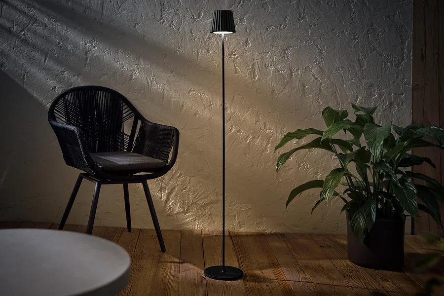 Černá LED stojací lampa na dobíjení pro venkovní použití stojí vedle černého proutěného křesla a rostliny v květináči.