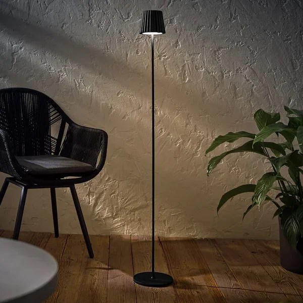Černá LED stojací lampa na dobíjení pro venkovní použití stojí vedle černého proutěného křesla a rostliny v květináči.