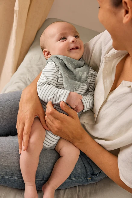 Matka drží usmívající se dítě, které má na sobě rostoucí body a mušelínový šátek.