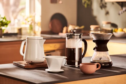 Na stole stojí bílý džbán, french press, překapávač a malý hrnek.