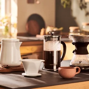 Na stole stojí bílý džbán, french press, překapávač a malý hrnek.