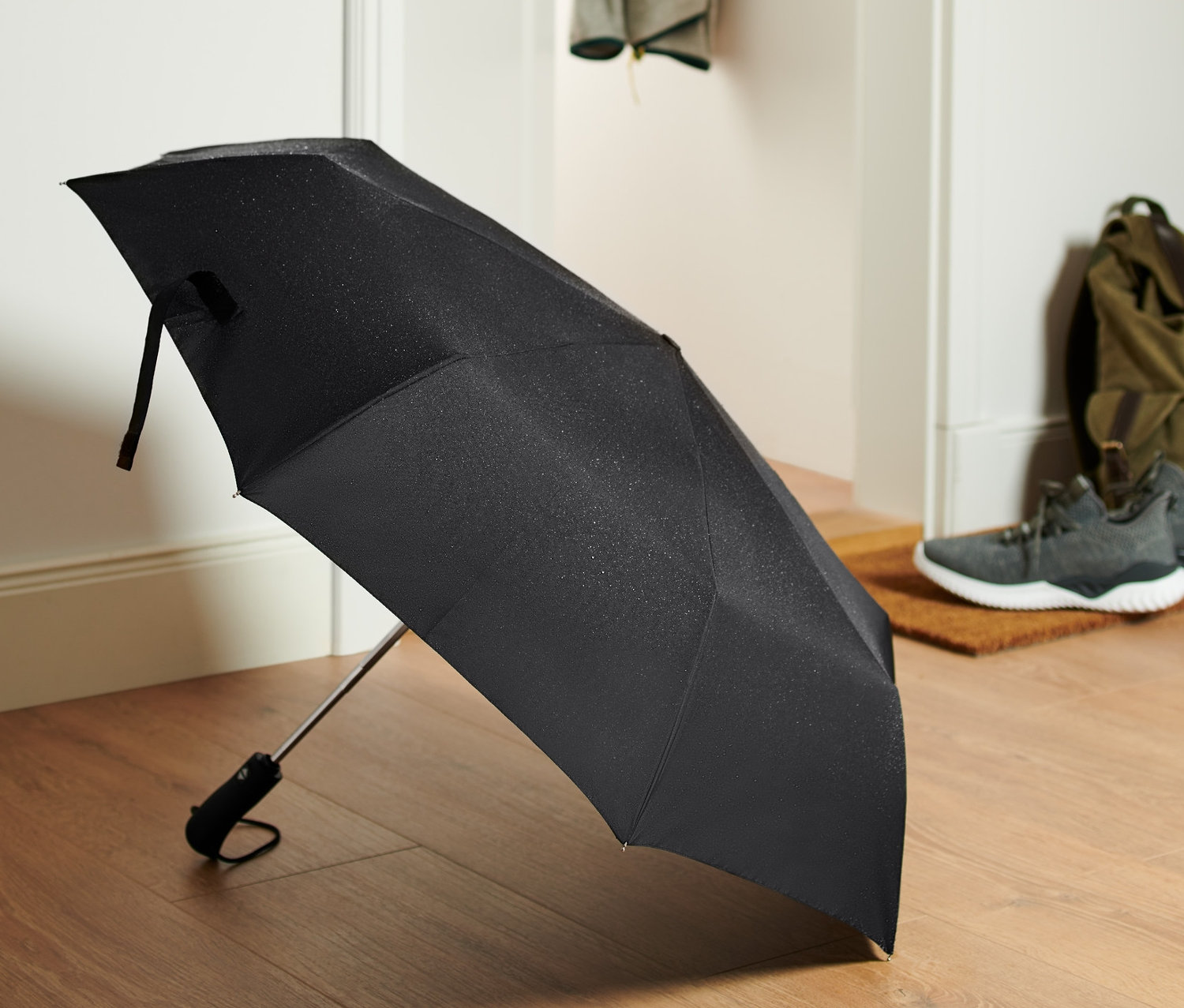 Černý automatický skládací deštník leží na dřevěné podlaze u dveří.