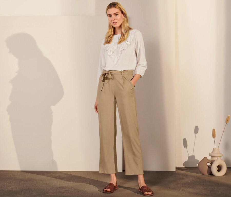 Žena pózuje v béžových kalhotách se sámky a bílé halence.