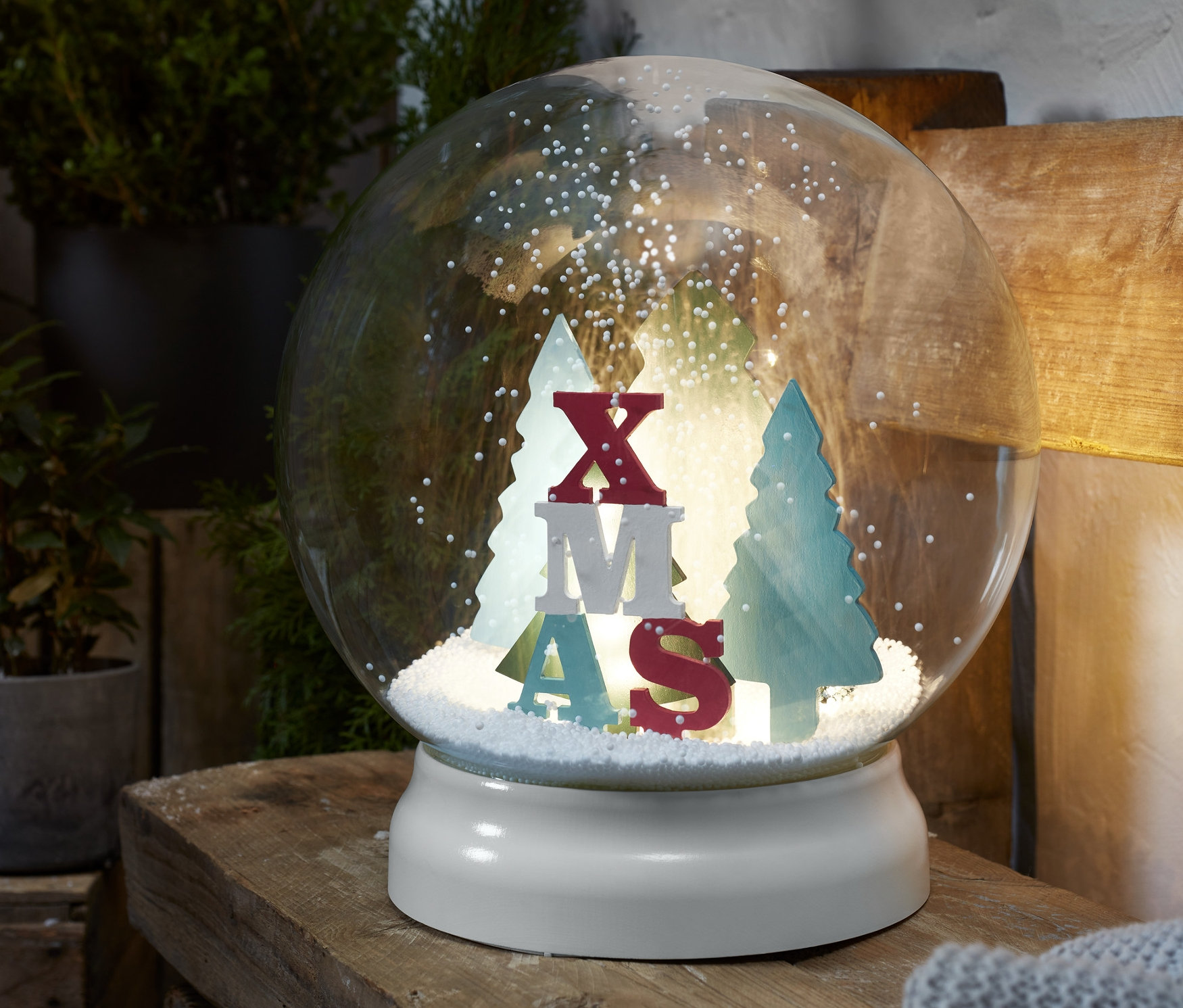 Venkovní sněžítko s LED osvětlením s nápisem XMAS a stromečky uvnitř, umístěné na dřevěném povrchu.
