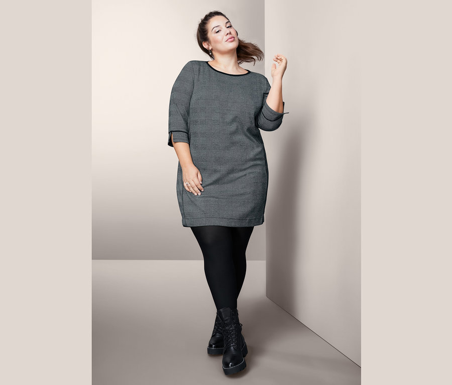 Žena pózuje v šatech z teplákoviny s 3/4 rukávy, černých punčochách a černých botách.