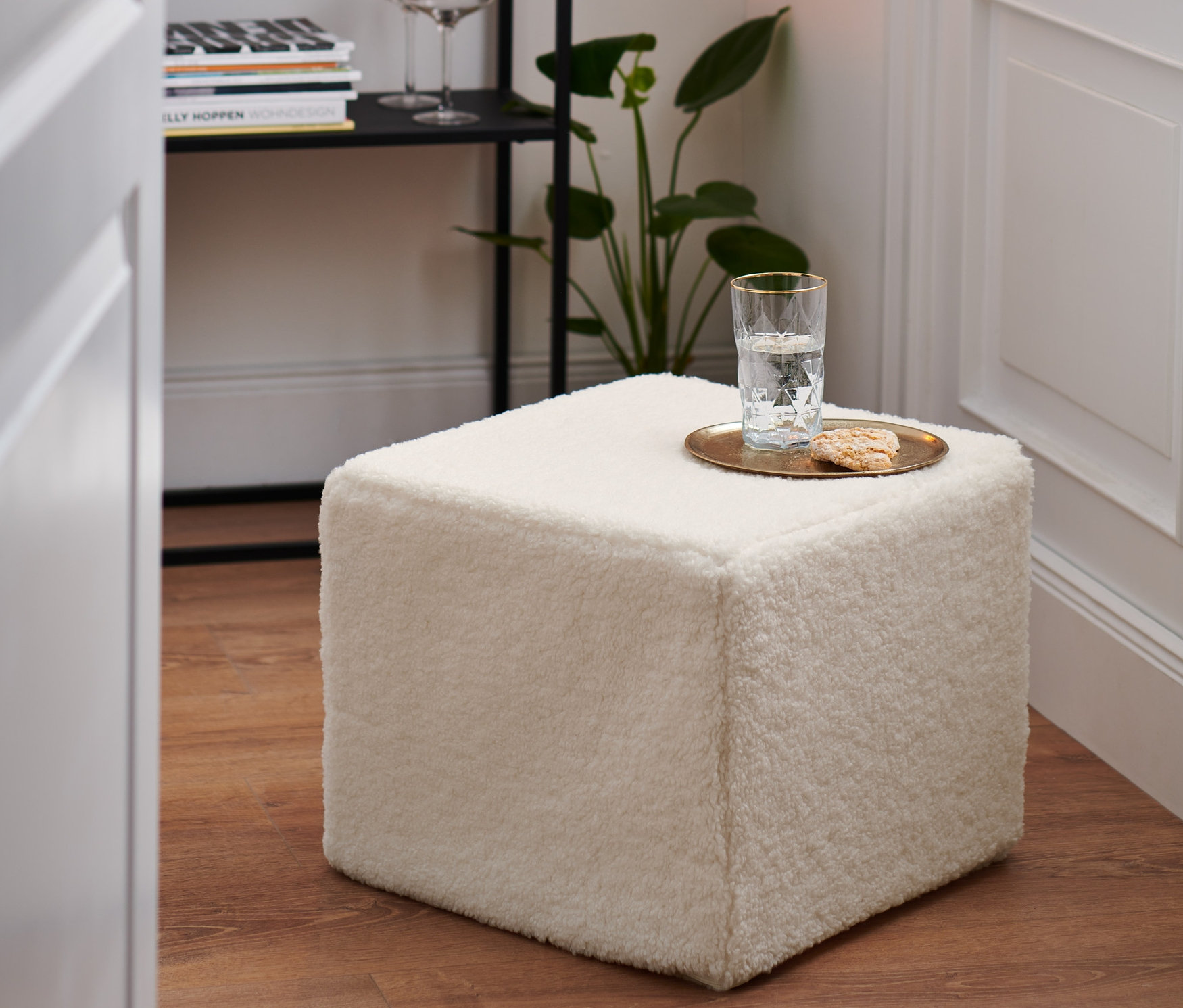 Bílý taburet stojí na dřevěné podlaze. Na taburetu je sklenice vody a sušenka.