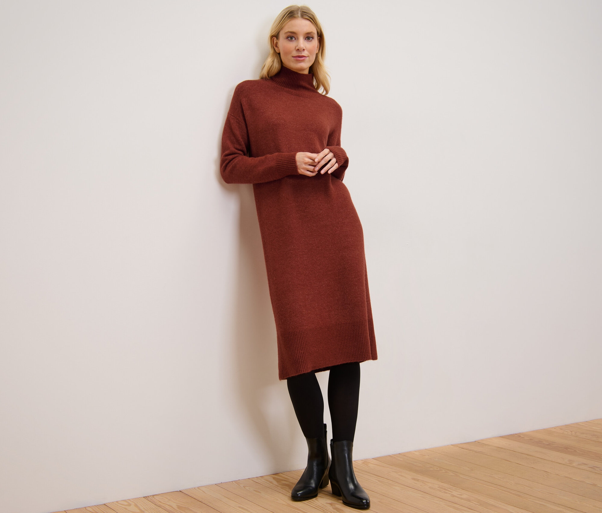 Žena opřená o zeď má na sobě pletené šaty s vlnou, černé punčochy a boty.