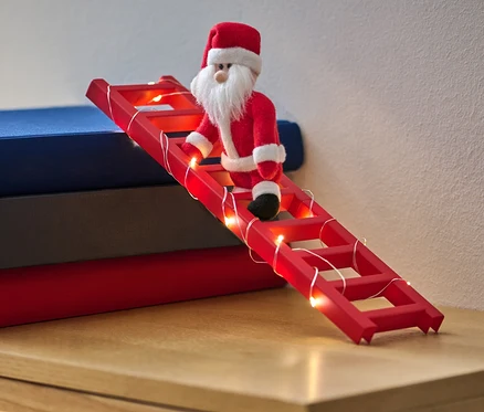 Santa Claus panenka leze po červeném žebříku s LED světelným řetězem, který je opřený o hromadu knih.