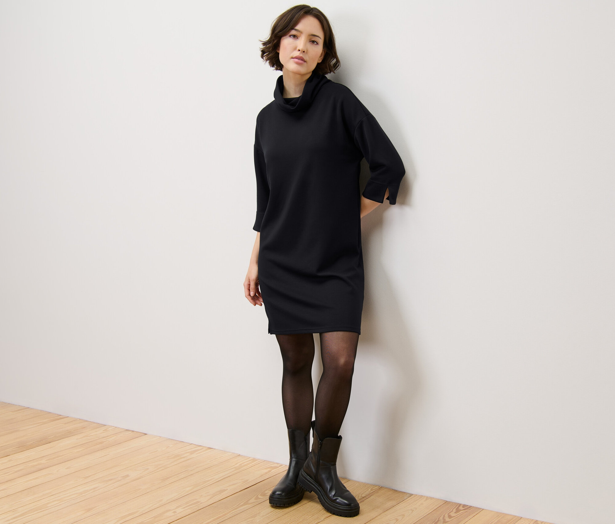 Žena stojící u zdi má na sobě teplákové šaty z modalové směsi, černé punčochy a černé boty.