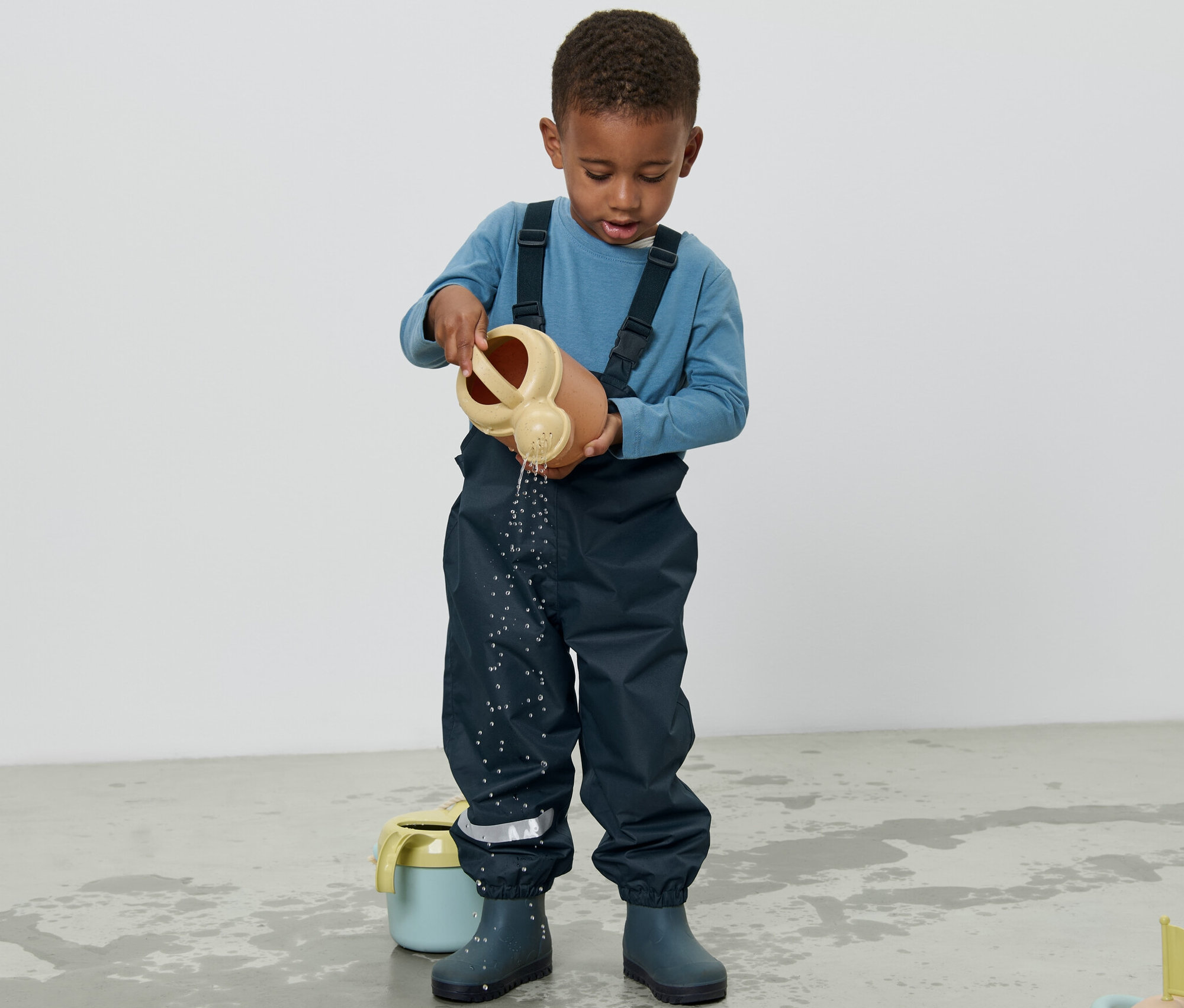 Chlapec v dětských kalhotách do deště lije vodu z konev na zem.