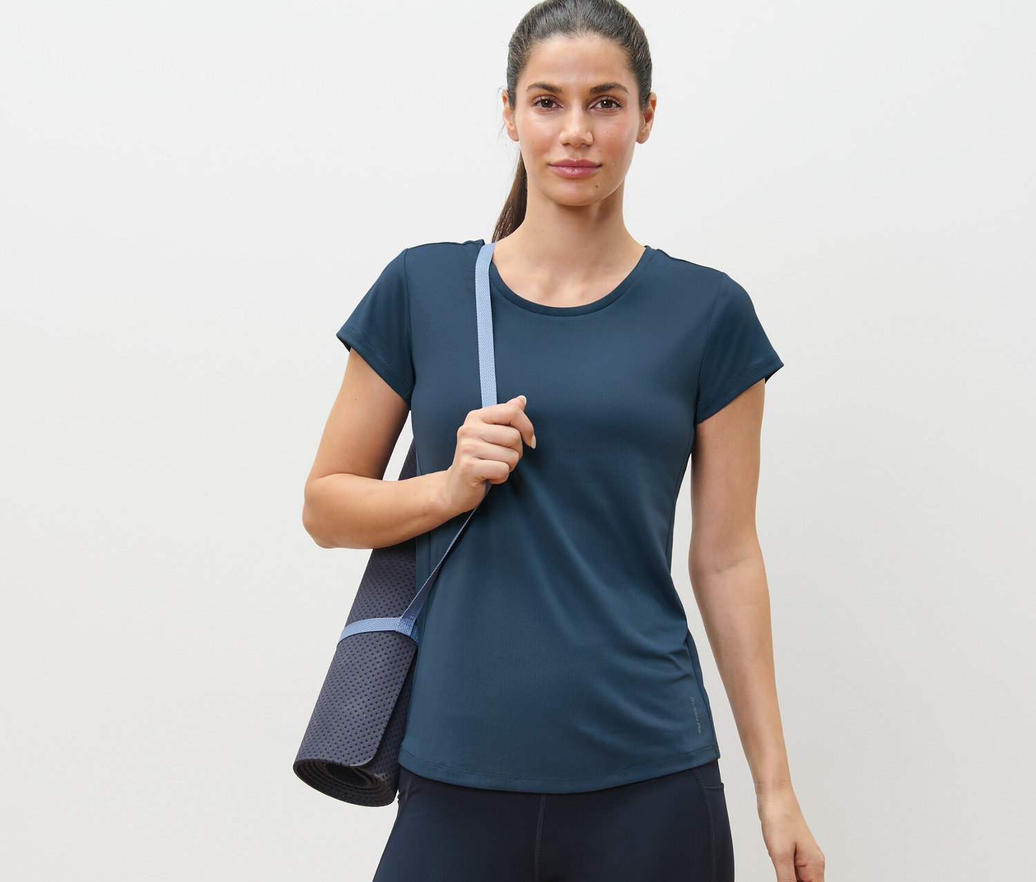 Žena má na sobě modré funkční tričko a nese podložku na cvičení.