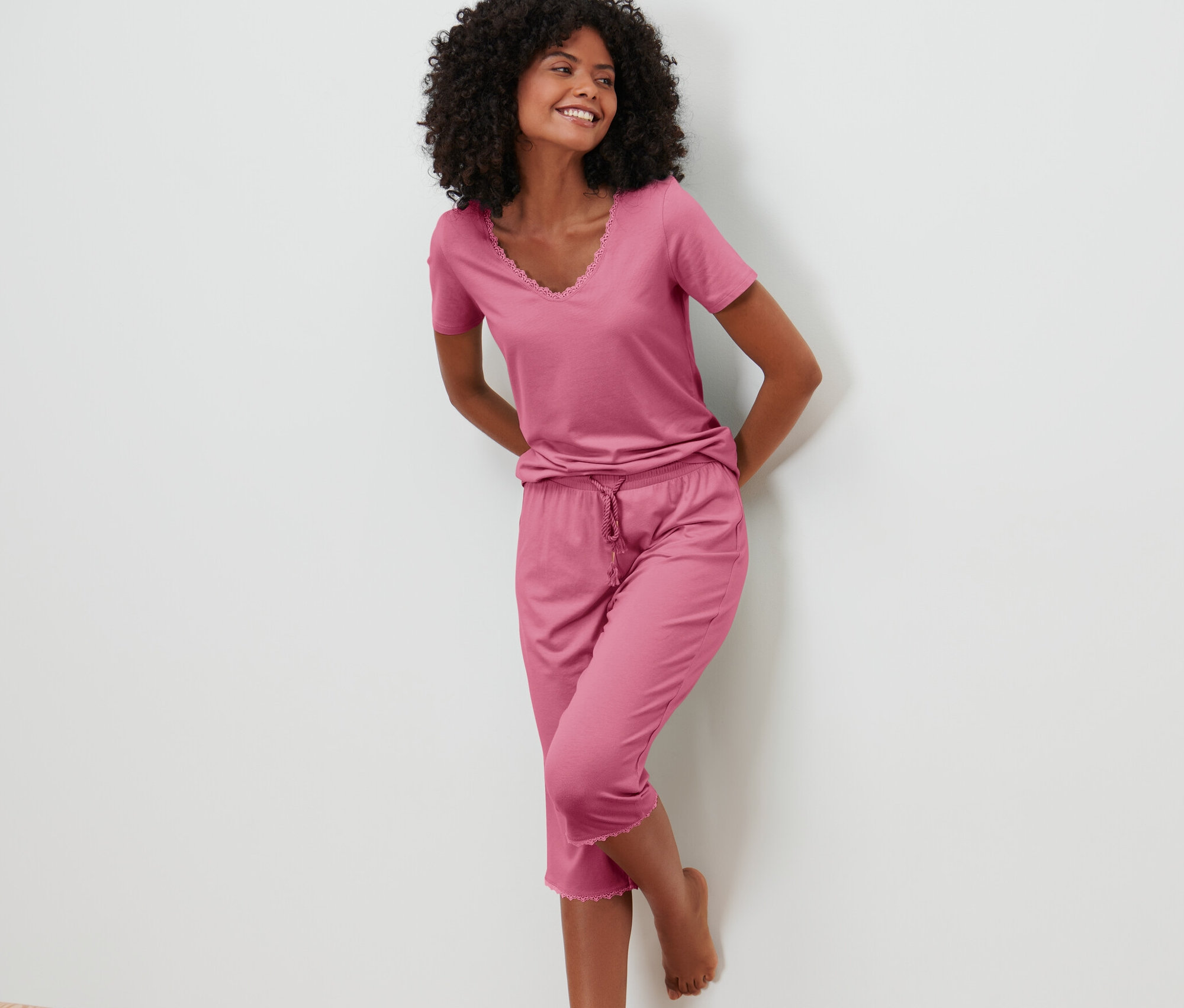 Žena se usmívá a opírá se o stěnu v růžové 3/4 pyžamové soupravě.