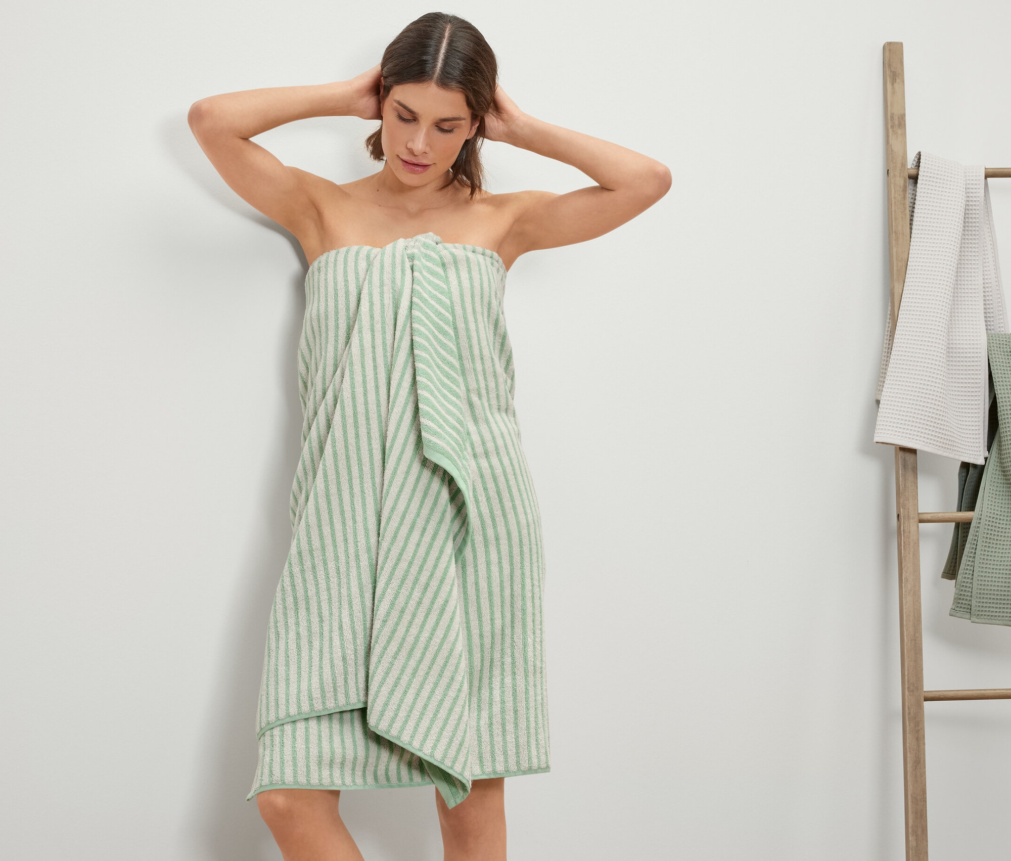Žena se zabalila do XL osušky s proužky. V pozadí je žebřík s dalšími ručníky.
