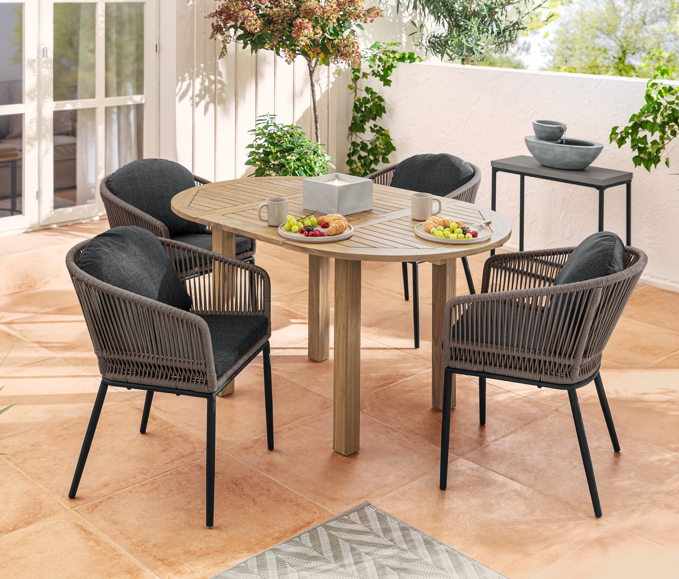 Čtyři jídelní křesla s kulatým opěradlem a textilním výpletem kolem kulatého rozkládacího stolu s jídlem a pitím na terase. V pozadí solární zahradní fontána a sada konzolových stolků.