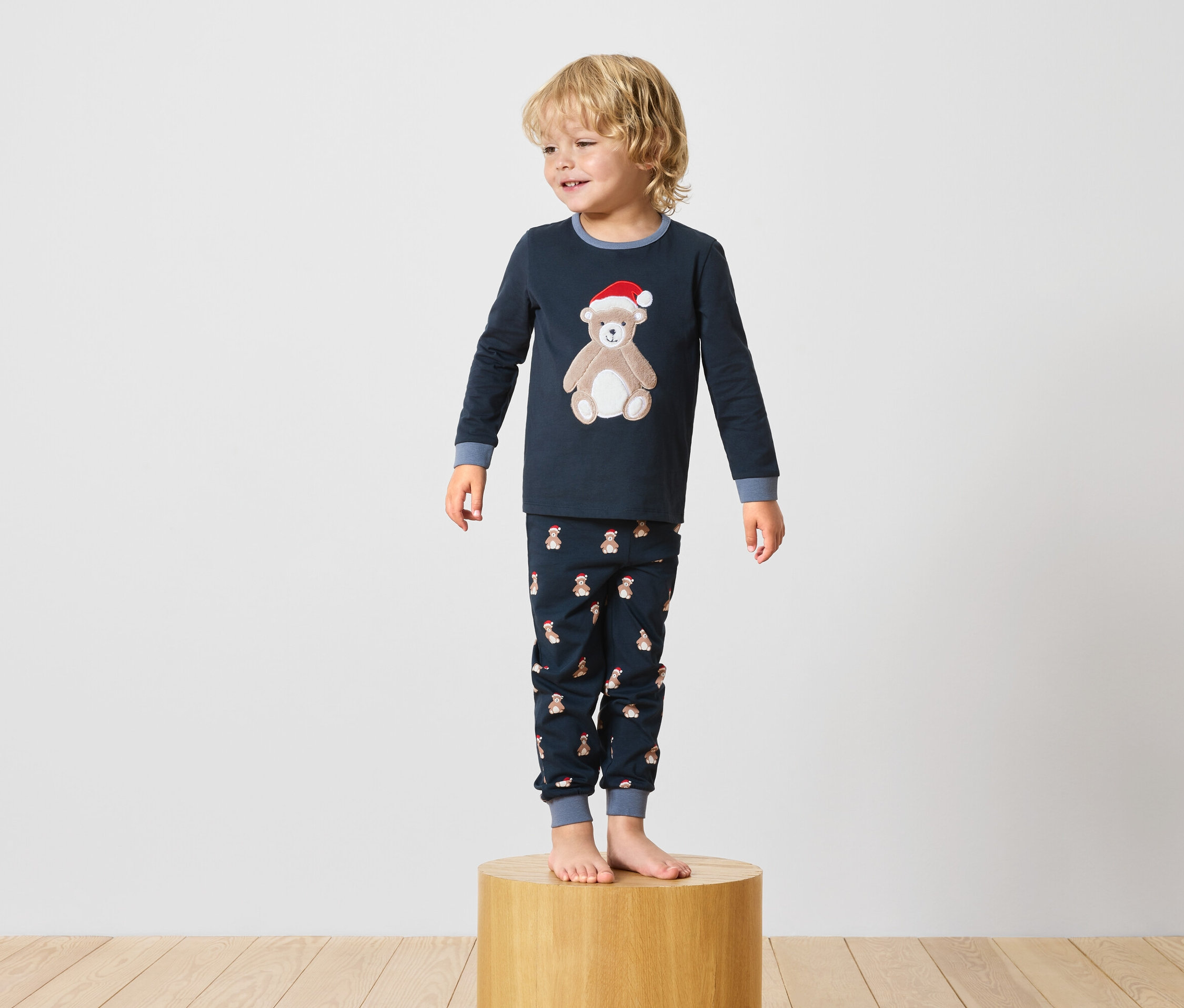 Chlapec stojí na dřevěném podstavci a má na sobě tmavě modré pyžamo s motivem medvídka.