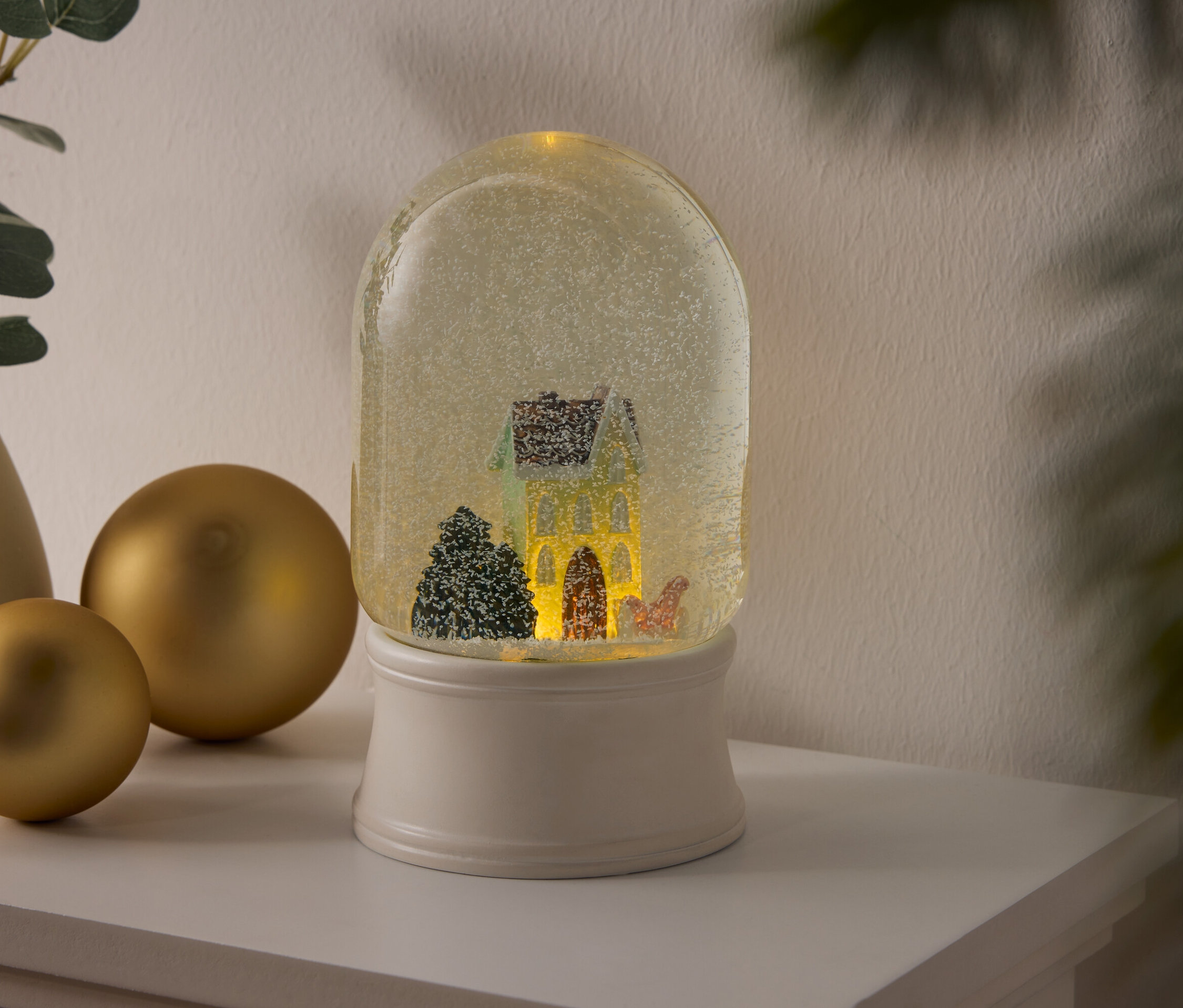 Sněhová koule s domem a stromy na bílém podstavci stojí na polici vedle zlatých koulí a vázy s rostlinami.