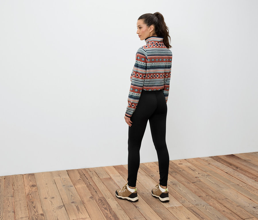 Žena s hnědými vlasy staženými do culíku stojí zády, má na sobě vzorovaný top s dlouhým rukávem, černé legíny a hnědé boty.