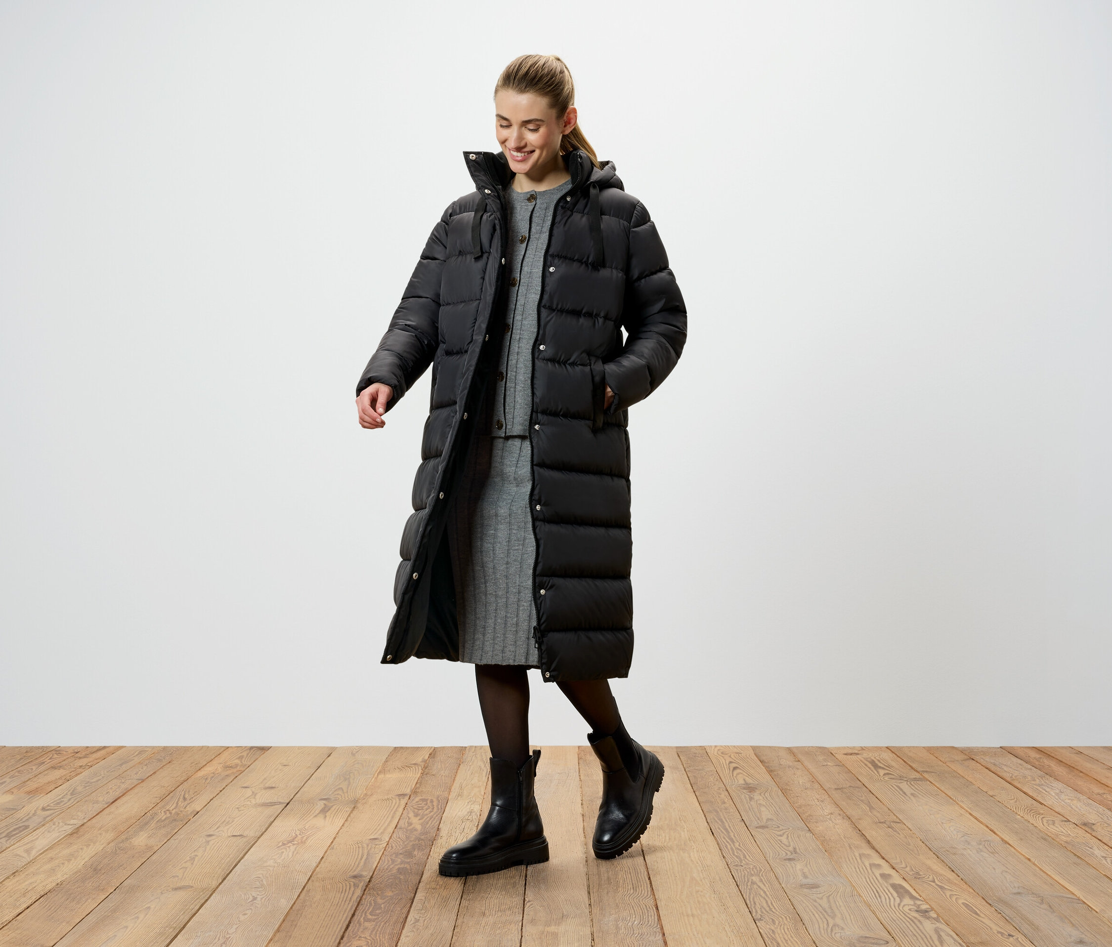 Žena v dlouhé černé péřové bundě, šedých pletených šatech a černých botách stojí na dřevěné podlaze.